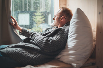 Portrait of thinking sad Middle-aged man in eyeglasses dressed open cardigan lying on cozy bed next...