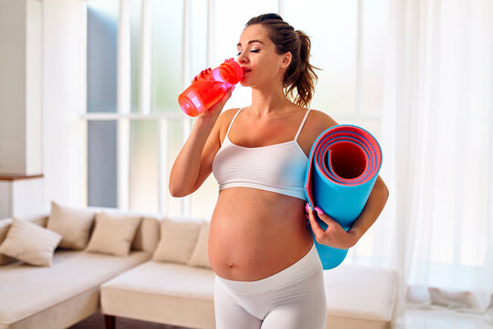 Young Pregnant Woman In Sportswear Holds A Rubber Mat And Drinks Water From A Bottle In The Living Room. Sports And A Healthy Lifestyle During Pregnancy.