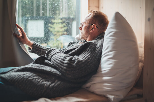 Portrait of thinking sad Middle-aged man in eyeglasses dressed open cardigan lying on cozy bed next to window looking out street through raindrop glass. Mental health and autumn weather concept image