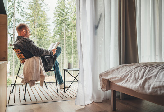 Alone man sitting in chair on country house balcony and enjoying forest view, tea cup, fresh air and bestseller novel thick book. Cozy bedroom interior back view. Reading or education concept image
