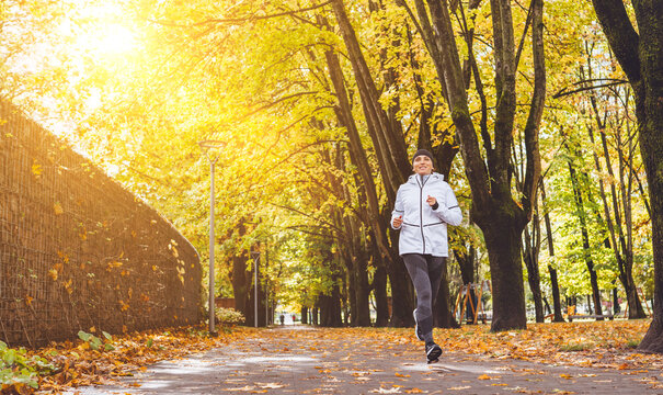 Fit athletic woman have a jogging in the autumnal city park. Young fitness female smiling and running by the footway covered with yellow leaves. Active running people concept image.