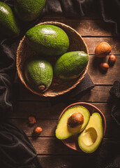 Some open and close ripe avocados in wooden bowls on a dark brown wooden table with black patterned and avocado seeds around. Dark food.