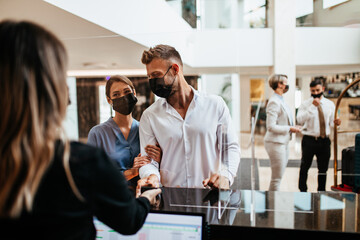 Young couple with protective face masks at the reception of a hotel checking in. Business trip, Coronavirus, Covid-19, safe travel concept.