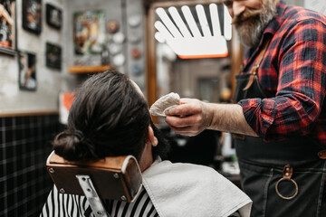 A barber trims a young guy's beard in his barber shop.