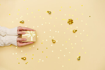 Female hands holding merry christmas gift box on yellow decorated holiday background, woman preparing surprise, new year winter merry holiday golden confetti from stars, real holiday concept, top view