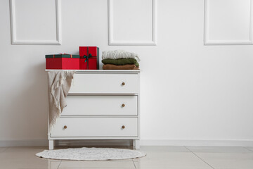 Chest of drawers with Christmas gift boxes and warm clothes near light wall