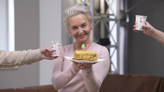 Portrait of happy confident beautiful Caucasian woman blowing out candles on 45 birthday smiling looking at camera as female hands appearing with paper glasses. Slim gorgeous lady posing indoors