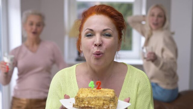 Joyful redhead beautiful woman with birthday cake blowing out 42 years candles smiling looking at camera as friends cheering. Portrait of cheerful confident happy lady posing at home celebrating