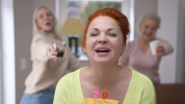 Front view happy woman blowing out candles on cake making wish looking at camera smiling. Portrait of excited beautiful Caucasian lady celebrating 50 birthday at home with friends posing