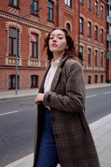 A wayward, proud young woman walks down a city street. Plaid coat, curly red hair