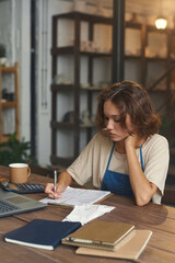 Busy female owner sitting at wooden table and making notes in papers while working with finances in pottery shop
