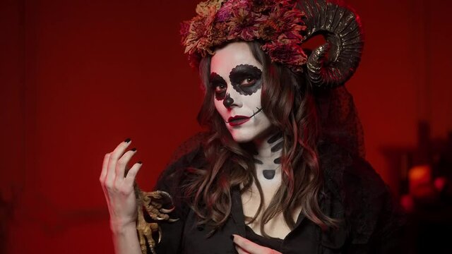 woman in catrina calavera style makeup on spanish day of the dead looks at the camera in a dark room with red lighting. Halloween witch costume with horns and dried flowers