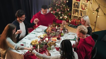 Friends holding hands and praying before dinner, new year tradition, sitting at dining table. International young people eating delicious food during christmas party at home. 
