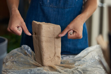 Close-up of unrecognizable potter in denim apron cutting clay with cut-off wire in workshop