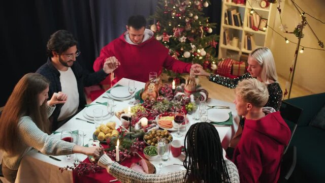 Friends holding hands and praying before dinner, new year tradition, sitting at dining table. International young people eating delicious food during christmas party at home. 