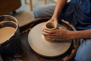 Close-up of unrecognizable potter sitting at pottery wheel and creating ceramic pot in workshop