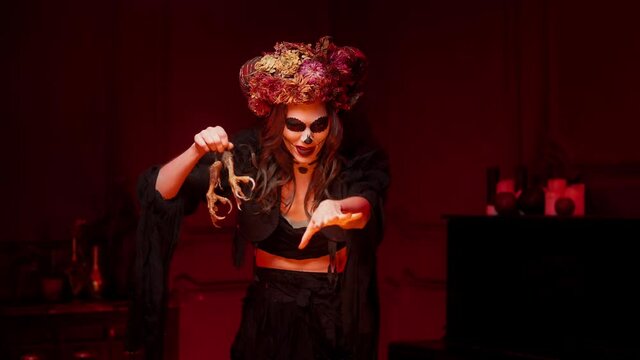 a sorceress in the style of a calavera casts a spell and throws bird paws with claws into the fire. Halloween Skull Makeup and Horns Costume for All Saints' Day and Spanish Day of the Dead