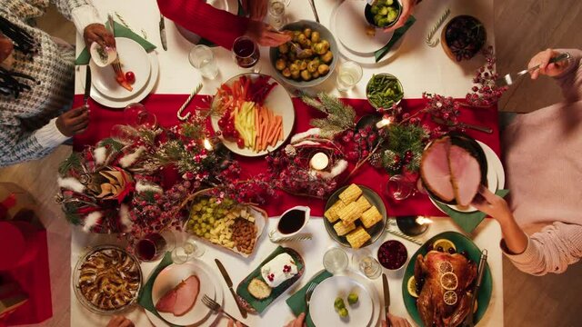 Friends celebrating new year, sitting at dining table top view. International young people eating delicious dishes, putting food on plates. Diverse students during christmas party at home. 