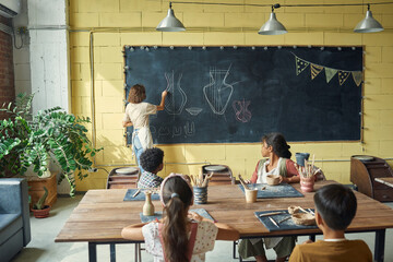 Rear view of pottery teacher drawing on blackboard while explaining multi-ethnic kids how to make...