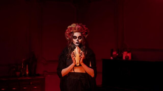 woman in image of witch for halloween with make-up in style of day of dead, horns, dried flowers on her head in a dark red room raises the skull of a wolf above her head, says a prayer by the fire