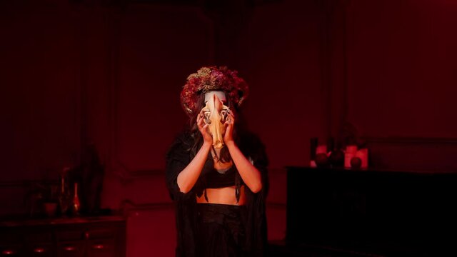 woman in rags halloween witch costume with calavera catrina makeup, horns, dried flowers on her head by the fire in a dark red room raises the skull of wolf to her chest, prays, utters spell, conjures