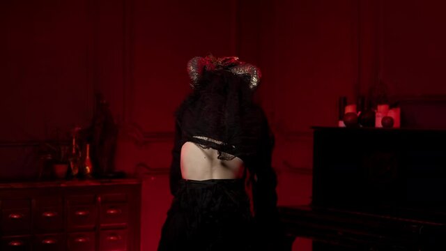 A female sorceress with horns, bird's paws spells over fire with her back to camera, turns sharply, looks, bewitches viewer. Halloween look in style of Spanish Day of Dead in dark room with red light