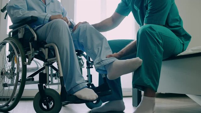 Male caregiver helping mature patient to recover injured leg, physiotherapy