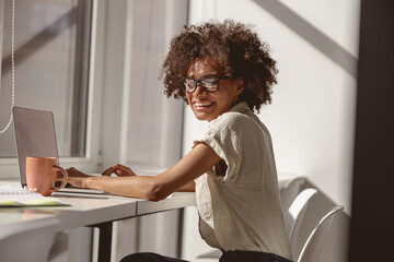 Fototapeta na wymiar Happy Afro American lady in glasses enjoying her job in the office while typing on laptop