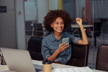 Happy Afro American lady sitting at the desk with laptop while holding smartphone