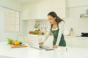 Portrait of happy smiling latin woman using a computer laptop notebook technology device, cooking food, working online at home in kitchen. People lifestyle. Household