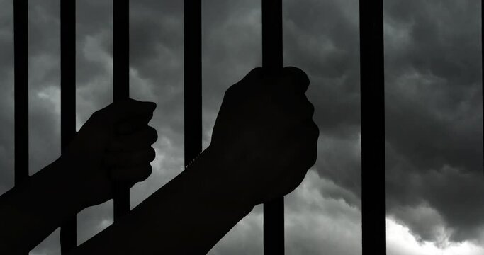 Prisoner hands holding prison cell bars with dark storm clouds background. 
