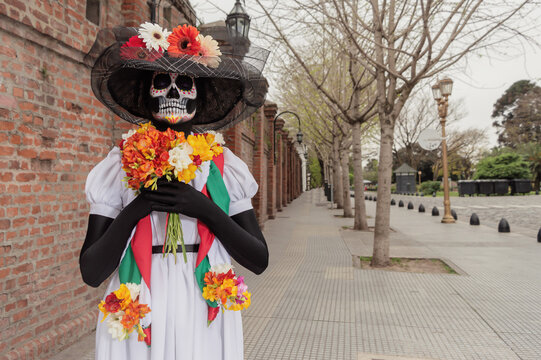 An adult woman with Catrina costume and make-up standing with her eyes closed and holding flowers outdoors