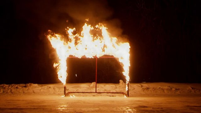 Carcass of hokey gate in large burning flame standing on empty ice arena with bank of snow at dark winter night