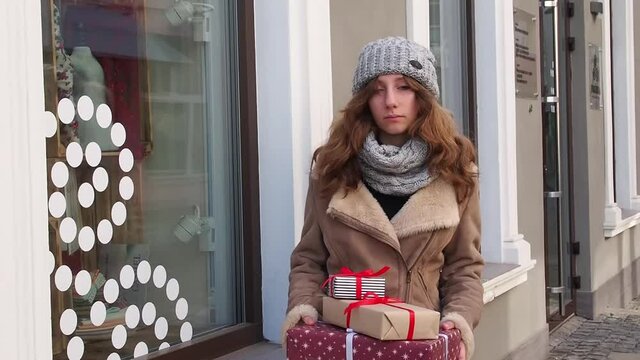 Teengirl in grey hat, scarf and brown jacket holding a set of christmas gifts near shops window. Happy young woman preparing holidays presents. Concept of shopping, giving and receiving gifts.