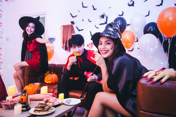 Group of young Asian friends in Halloween costumes and having fun with cocktail and snack at home party. Pretty woman wearing as a witch smiling and looking at the camera on foreground.