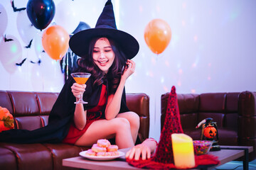 Smiling Asian woman in Halloween costumes having fun with cocktail drink and snacks at home. Pretty young girl wearing as witch smiling and enjoying and celebrating Halloween party.
