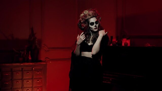 a witch in the form of Catrina with horns and dried flowers dances in a dark red room to the flashes of light. Make-up, halloween costume in the form of a painted skull with a sewn-up mouth