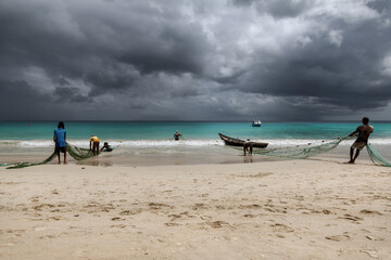 Fishermen on a beach pull nets from the sea in sandy beach with dramatic sky on Mahe Island,...
