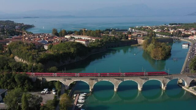 Red high-speed train on the bridge. Frecciarossa high-speed train passes an arched railway bridge over a river in the background Lake Garda Italy.