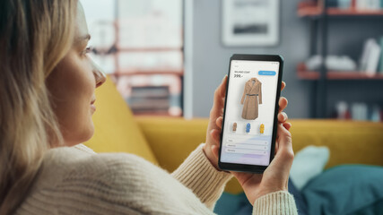 Beautiful Caucasian Female is Using Smartphone with Clothing Online Web Store to Choose and Buy Coat from New Collection. Female Surfing the Net and Lying on Couch Sofa at Home Living Room.