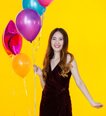 Portrait studio shot of Asian happy girl wears sexy dress standing posing smiling look at camera holding colorful helium air balloons in hand in celebration party in front of yellow wall background