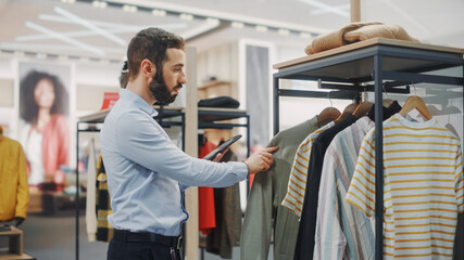 Modern Clothing Store: Male Visual Merchandising Professional Uses Digital Tablet Computer To...