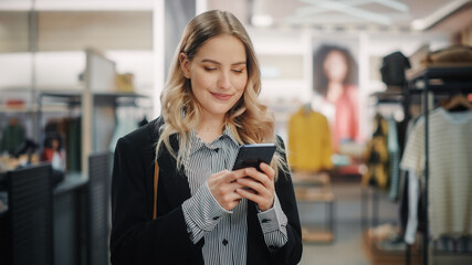 Beautiful Smiling Female Customer Shopping in Clothing Store, Using Smartphone, Browsing Online, Comparing on Internet, Choosing Stylish Clothes. Fashionable Shop, Colorful Brands, Sustainable Designs