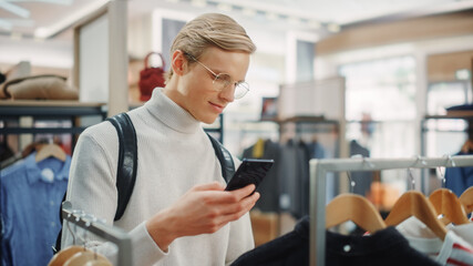 Handsome Male Customer Shopping in Clothing Store, Using Smartphone, Browsing Online, Comparing on...