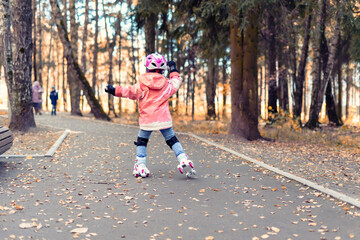 kid girl in a pink helmet rides in an autumn park on four-wheeled rollers. autumn outdoor activities