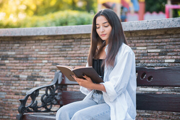 A young and beautiful Arab or Caucasian girl dressed in casual style is reading a book while sitting on a bench in the park. Knowledge and education concept.