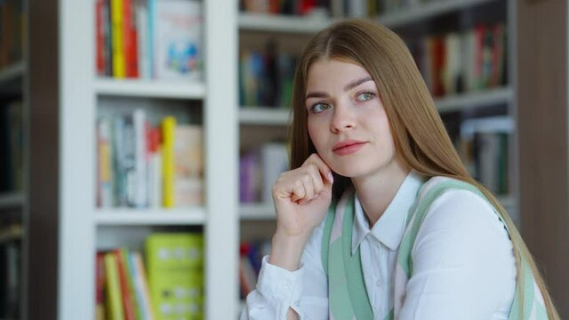 Young woman with long hair daydreaming against bookcases on blurred background. Tracking shot college student studying in library. Concept of education