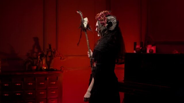 a voodoo woman walks with a staff with a skull through a dark fiery room, turns to face the camera, looks. Halloween skeleton style witch image with horns, sewn mouth