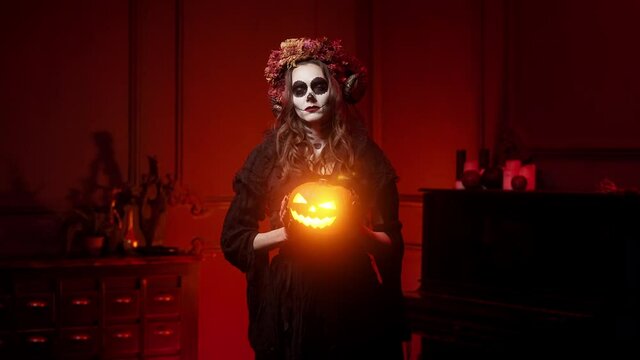 witch in form of calavera with horns, a sewn-up mouth on Halloween night with a jack-o-lantern in a dark burning room. Makeup, all saints' day costume in style of spanish day of dead