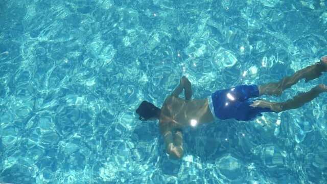 Brunet boy in shorts swimming underwater in a light-blue wading pool in slo-mo 